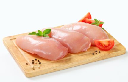 Three Chicken breast boneless 1 kg on a wooden cutting board, garnished with green herbs and accompanied by sliced tomatoes. Scatterings of peppercorns complete the scene against a plain white background. Smart Chicken