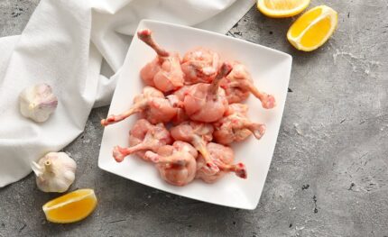 Chicken lollipop 1 kg placed on a white square plate on a gray textured backdrop, accompanied by halved garlic bulbs and two lemon wedges. A nearby white cloth adds to the setup. Smart Chicken