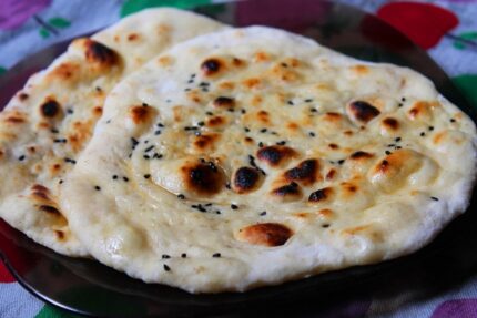A single Kulcha roti with a golden-brown surface, sprinkled with black sesame seeds, lies on a dark plate atop a colorful, patterned tablecloth. Smart Chicken