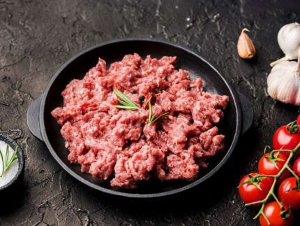 A black plate of Mutton keema 1 kg is garnished with rosemary and peppercorns, surrounded by cherry tomatoes, garlic bulbs, and a small bowl of salt on a dark textured surface. Smart Chicken
