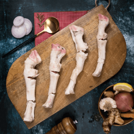 Four pieces of mutton paya on a wooden cutting board are surrounded by onions, a gold spoon, a red mat, a bowl with ginger and shallots, and a pepper grinder against a dark textured background. Smart Chicken