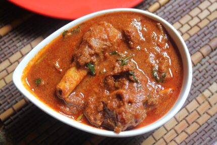 A white bowl of Mutton curry 4 Piece, showcasing vibrant red Indian curry with meat pieces and fresh coriander garnish, rests on a woven mat. Smart Chicken